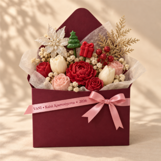 VANI Écrin Fleuri de Noël — χριστουγεννιάτικη ανθοσύνθεση από κερί σε μπορντό φάκελο, με κόκκινα και ιβουάρ λουλούδια, δεντράκι και δωράκι wax melts, λευκό αλεξανδρινό και ροζ κορδέλα VANI.